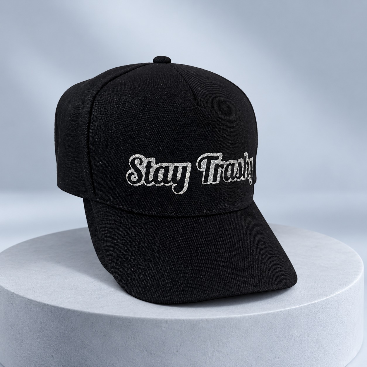 Black Stay Trashy hat with silver lettering on a pale blue-gray studio plinth